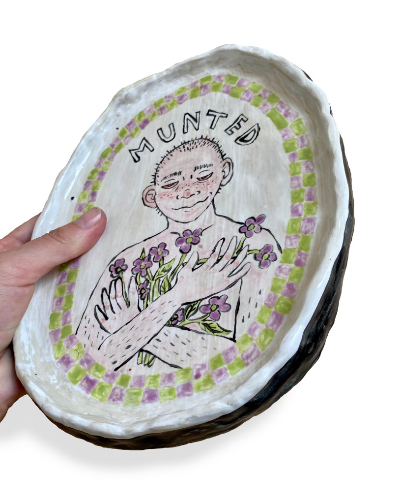 A picture of an oval ceramic plate with a depiction of a topless boy with a shaved head (Chuck Beans) holding a bouquet of violets to his chest and smiling. He is very pink and has freckles and bloodshot eyes that are half-closed. Above him reads the word "Munted". There is a border of green and purple checkers around the edge of the plate. A hand is tilting the plate so that the black edge of the plate can be seen.